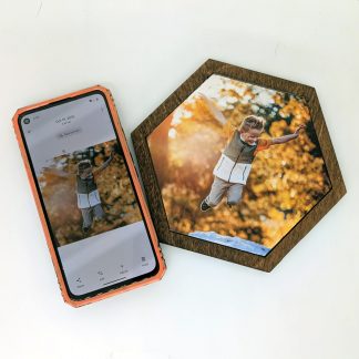 Smartphone displaying a child jumping in fall leaves next to a matching UV-printed custom hexagon wood tile in a dark walnut frame