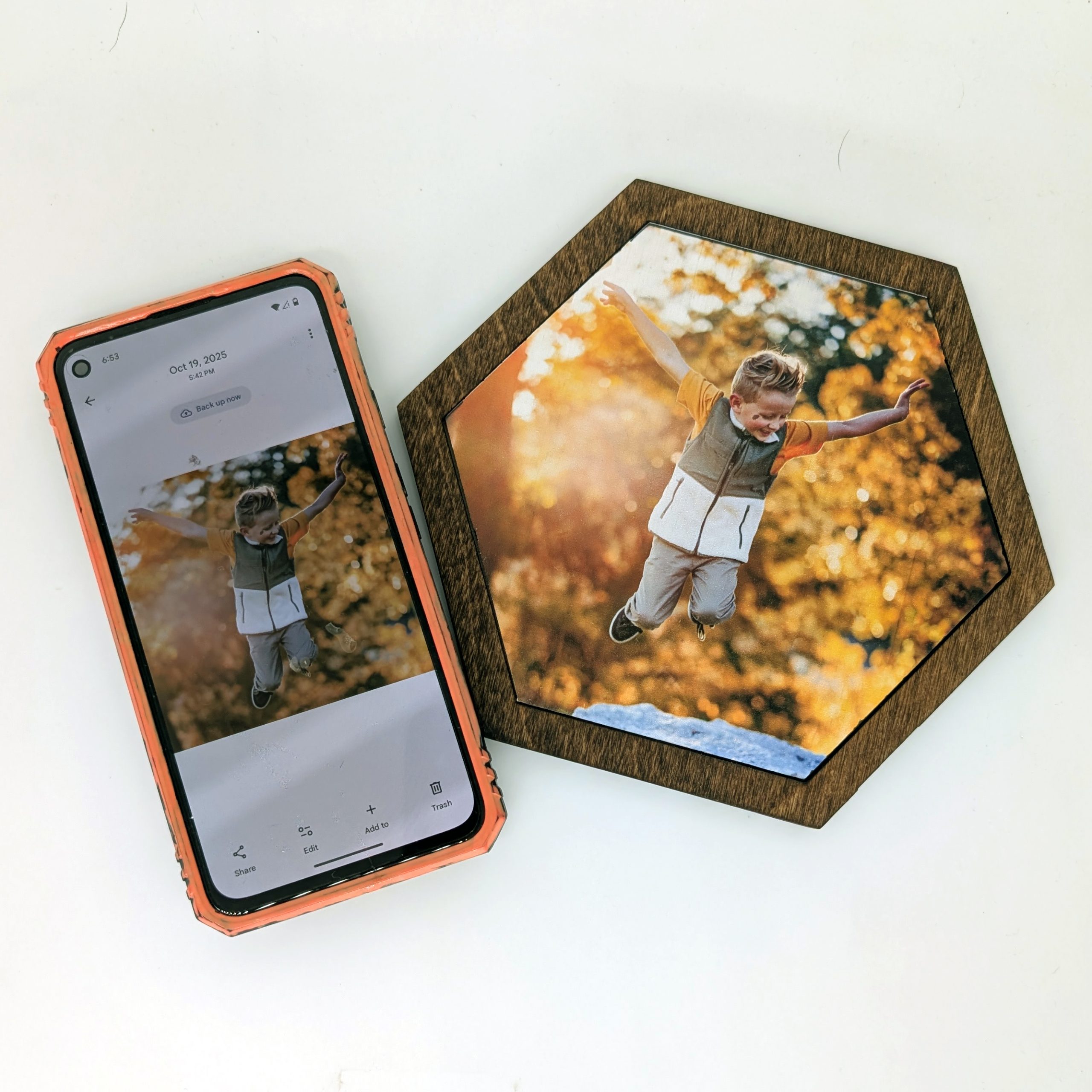 Smartphone displaying a child jumping in fall leaves next to a matching UV-printed custom hexagon wood tile in a dark walnut frame