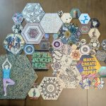 A large grouping of hex wall tiles in various sizes and designs laid out on a wood surface, showing henna patterns, pop art, vintage prints, and a yoga pose tile