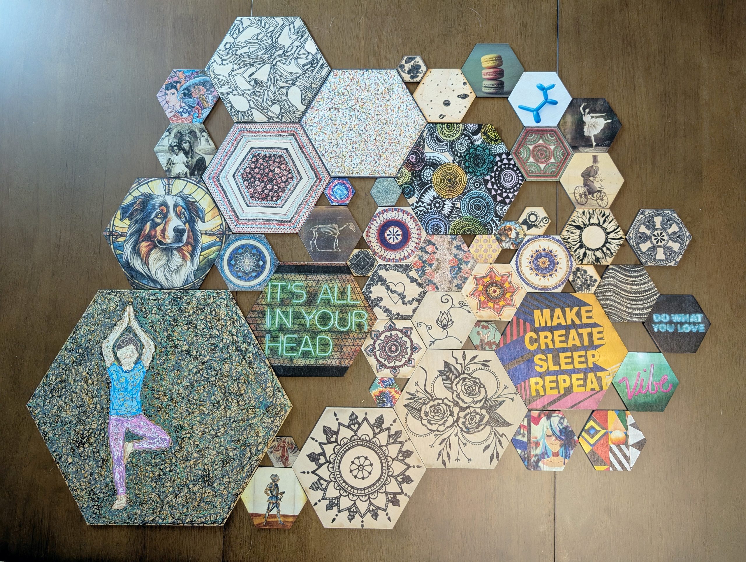 A large grouping of hex wall tiles in various sizes and designs laid out on a wood surface, showing henna patterns, pop art, vintage prints, and a yoga pose tile